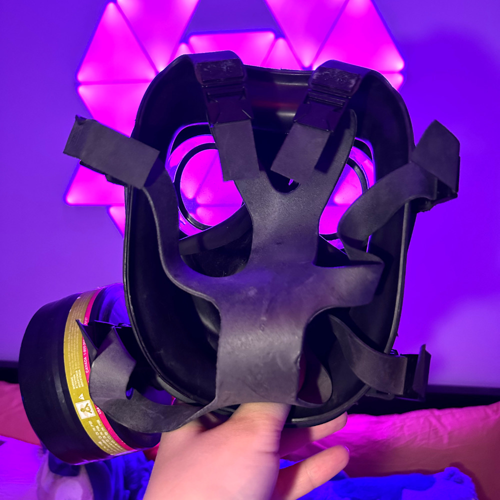 Rina holding a black S10 gas mask with the inside towards the camera. The head straps cover most of the inside, but you can see out of the clear lenses, at the purple cyber fox lights on the wall behind it. The ambient lighting is now dark purple